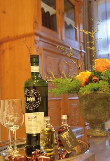 Grüne Whiskyflasche auf einem Tablett mit Whiskygläsern und Samples vor einem Blumengesteck und antikem Fichtenholzschrank.