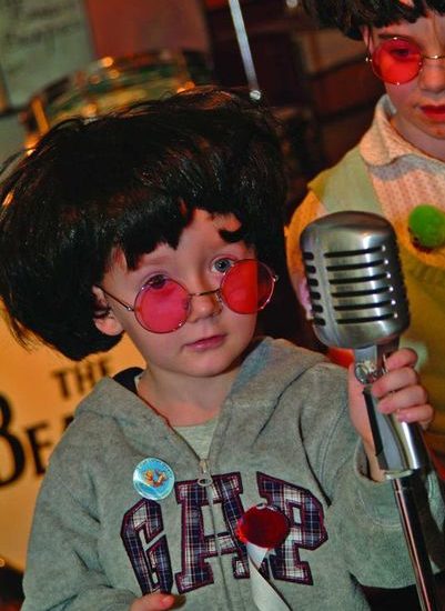 Ein kleiner Junge mit grauer Jacke, roter John Lennon Brille und schwarzer Perücke hält ein altes Mikrofon in der Hand.