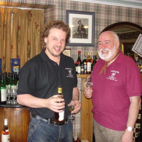 Zwei Männer stehen mit Whiskyglas in der Hand in einem mit Tartan dekoriertem Raum mit zahlreichen Whiskyflaschen.