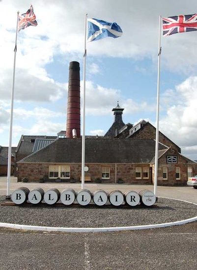 Vor dem Bruchsteingebäuder der Destillerie liegen Fässer mit den Buchstaben Balblair, flankiert von drei Fahnenmasten mit britischen und schottischen Flaggen.