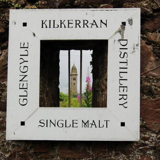 Quadratisches Schild mit Beschriftung Kilkerran Glengyle Single Malt Distillery, durch das man durch eine Mauer auf einen Kirchturm schaut.