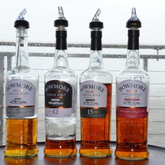 Vier Flaschen von Bowmore vor einer regennassen Scheibe und grauem Meer.