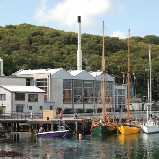 Caol Ila Distillery mit einem großen Gebäude mit Glasfenstern, hinter denen Kupferbrennblasen zu erkennen sind. Davor liegen bunte Segelboot am Steg.