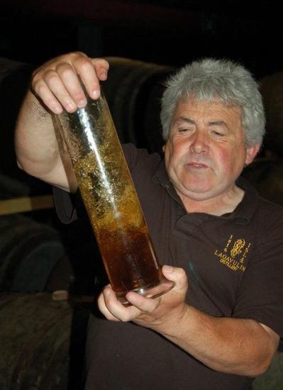 Mann mit schwarzem Polohemd der Lagavulin Distillery schüttelt ein Glasgefäß mit Whisky.