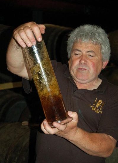 Mann mit schwarzem Polohemd der Lagavulin Distillery schüttelt ein Glasgefäß mit Whisky.