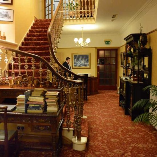 Eingangsbereich eines Hotels dessen große Treppe mit antikem Metallgeländer links dominiert. Vorne ein Tisch mit Büchern.