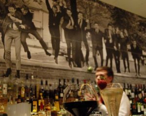 Bar mit Cocktailgläsern und im Hintergrund Flaschen vor einem Wandbild mit feiernden Männern in schwarzen Anzügen.