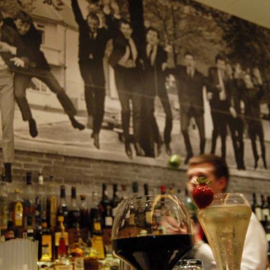 Bar mit Cocktailgläsern und im Hintergrund Flaschen vor einem Wandbild mit feiernden Männern in schwarzen Anzügen.