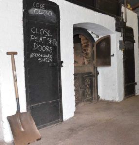 Eine große Schaufel steht vor einer weißen Wand mit schwarzer hoher Eisentür. Daneben ist eine alte Feuerstelle für Torffeuer.