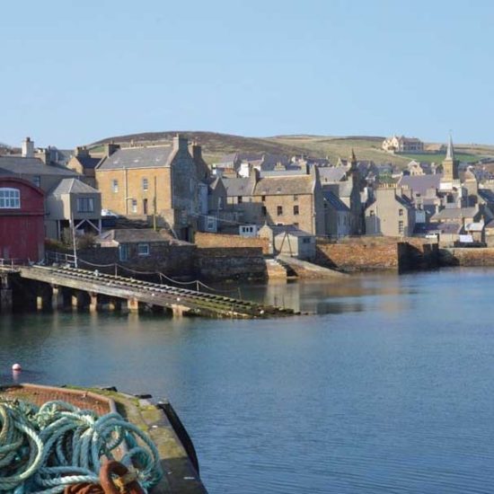 Nordische Hafenszene mit graubraunen Steingebäuden und einem roten Bootsschuppen, von dem eine Rampe ins Meer führt. Im Vordergrund hellblaue Seile,