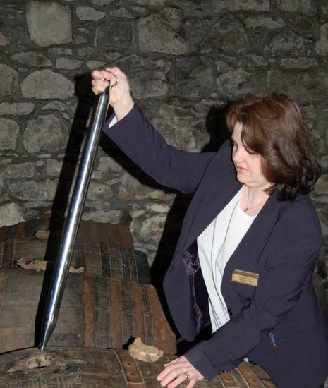 Eine Frau mit blauem Sakko zieht mit einer Valinch eine Probe aus einem Whiskyfass.