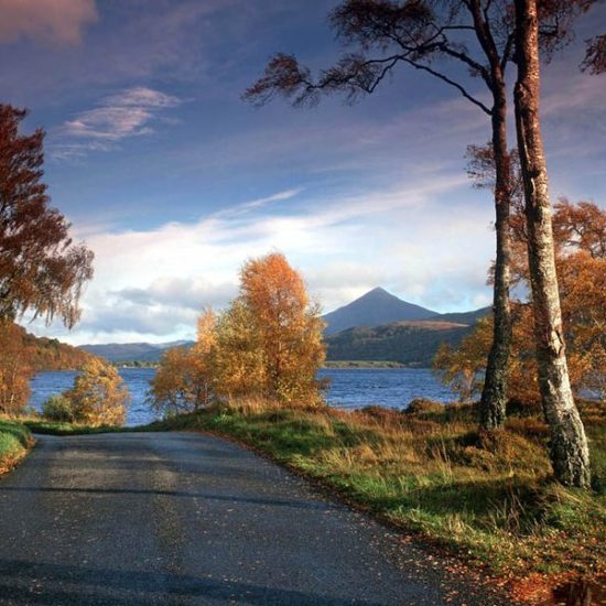 Eine schmale geteerte Straße führt zu einem See, der vor einem spitzen Bergkegel in der Herbstsonne glänzt. Die Bäume haben Herbstfärbung.