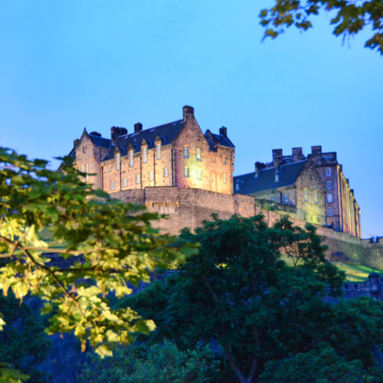 Das beleuchtete Edinburgh Castle wird im blauen Abendlicht im Vordergrund von Ästen gerahmt.