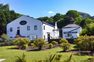 Zwei weiße Gebäude der Glengoyne Distillery liegen in einer gepflegten Gartenanlage vor dem Berg Dumgoyne.