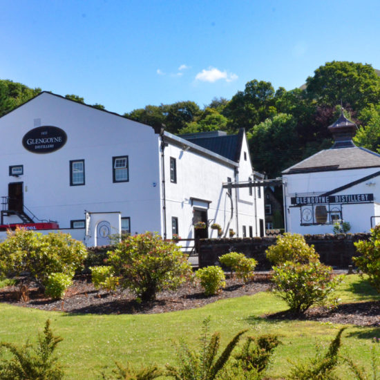 Zwei weiße Gebäude der Glengoyne Distillery liegen in einer gepflegten Gartenanlage vor dem Berg Dumgoyne.