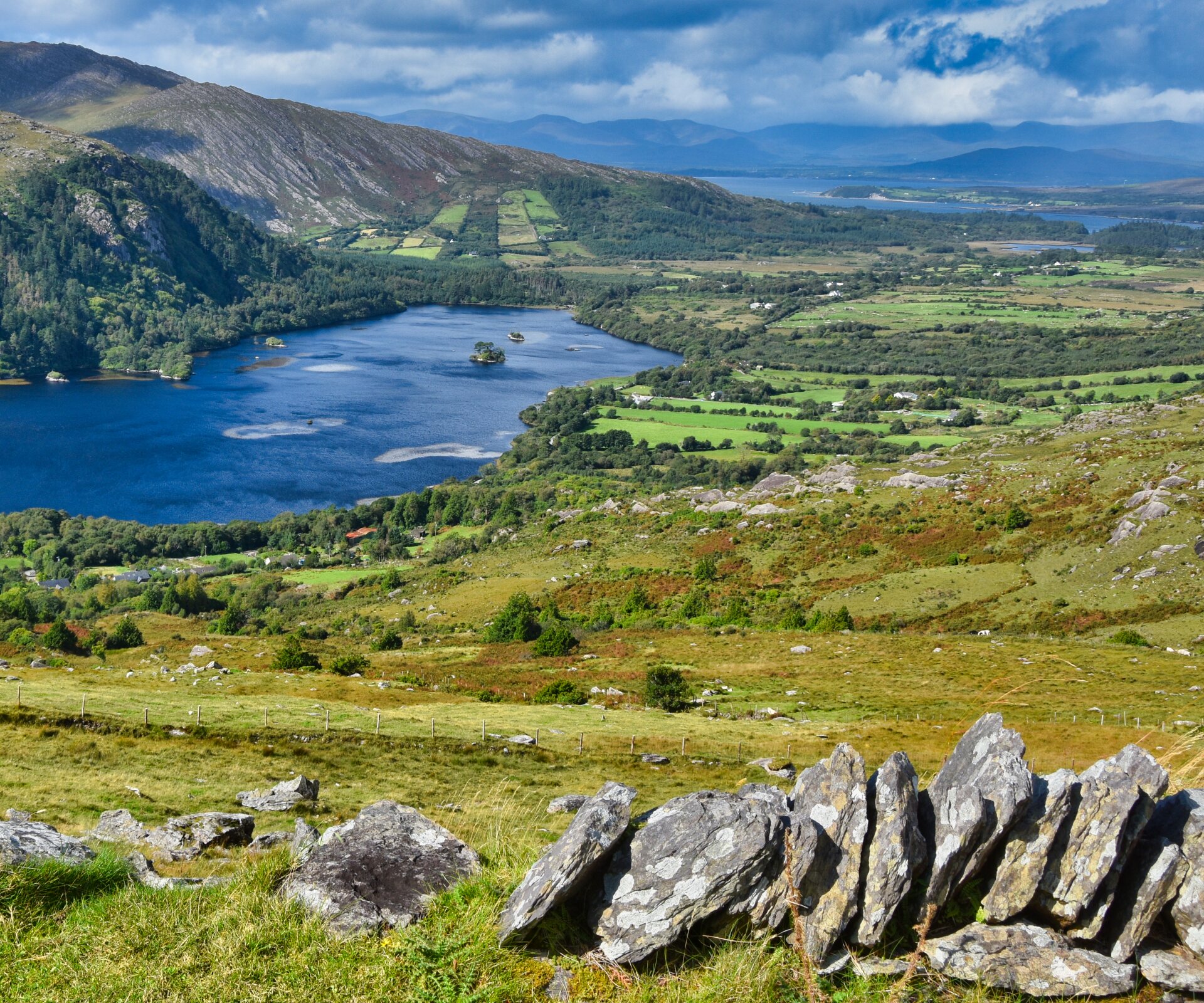 Blick von einer Passtraße in Irland weit über die Landschaft hinweg. Links ein Bergsee, davor Weiden und im Hintergrund Berge.