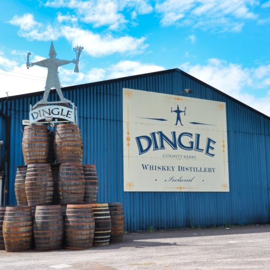 Blaue Lagerhalle mit einem Firmenschild der Dingle Distillery, vor der eine Turm aus mehreren großen Whiskyfässern aufgebaut wurde. Darauf steht eine Metallfigur.