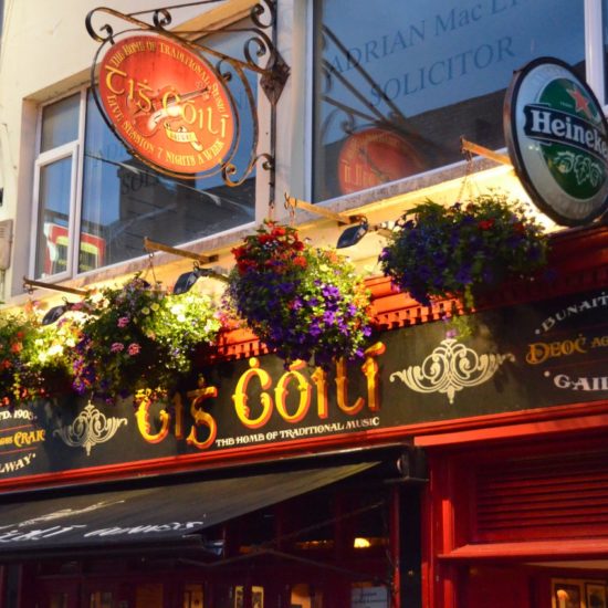 Werbeschilder eines irischen Musikpubs, an dessen Fassade auch Blumenkörbe hängen.