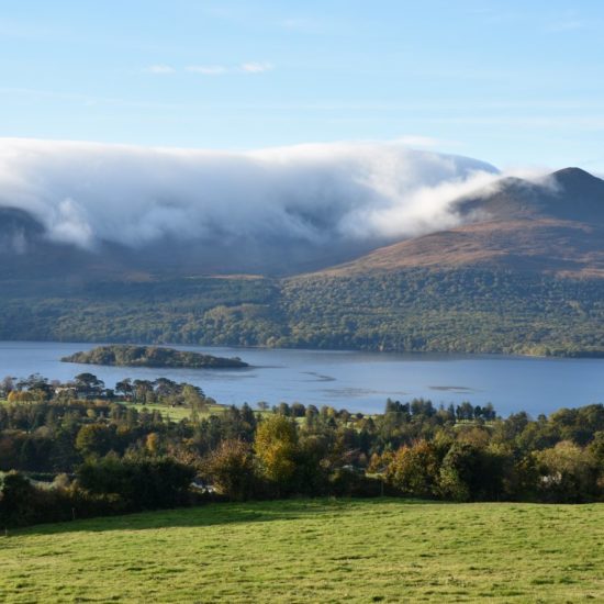 Herbstliches Panorama mit einem See. Im Vordergrund eine Wiese, im Hintergrund Berge über die sich eine Wolkendecke legt.