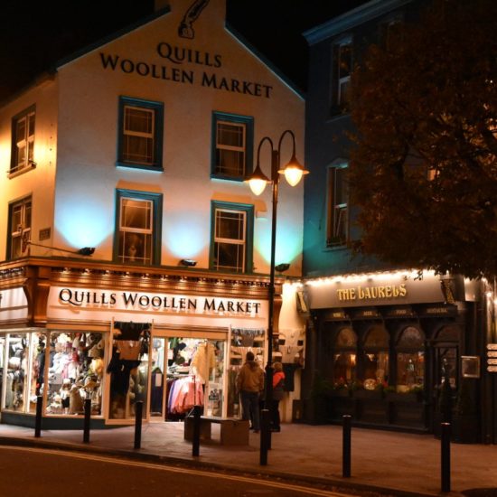 Nächstliche Straßenszene mit einem beleuchteten Wollgeschäft an einem kleinen Platz in Killarney.