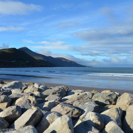 Küstenschutz durch große Steinbrocken an einem breiten Sandstrand mit den Bergen des Ring of Kerry im Hintergrund.