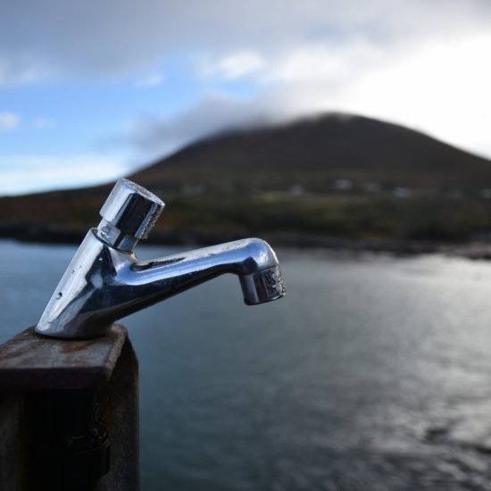 Wasserhahn auf einem Holzblock,vor dem Hintergrund einer Meeresbucht, die durch den Wasserhahn gefüllt zu werden scheint.