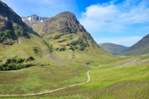 Ein Wanderweg windet sich durch ein grünes, von hohen, fast waldlosen Bergen gerahmten Tal von Glen Coe in Schottland.