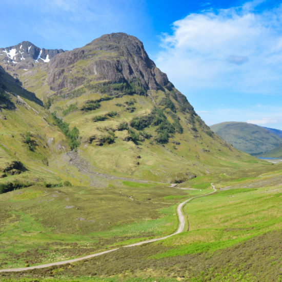Ein Wanderweg windet sich durch ein grünes, von hohen, fast waldlosen Bergen gerahmten Tal von Glen Coe in Schottland.