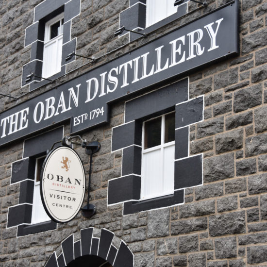 Dunkle Steinfassade mit schwarzen Fensterrahmen und Beschriftung The Oban Distillery.