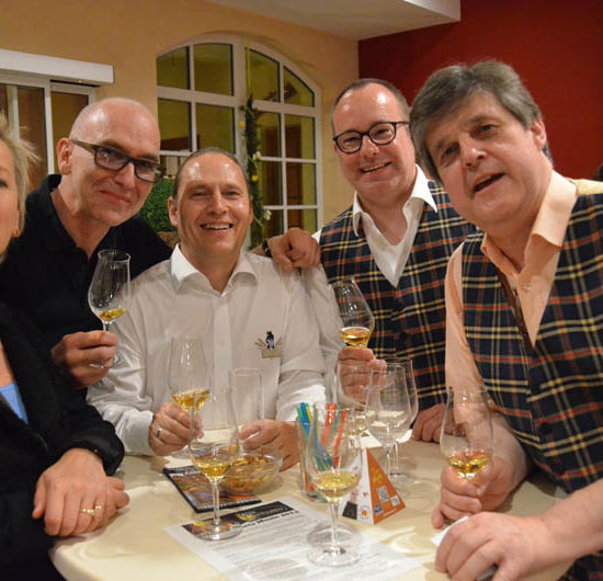 Vier Männer und eine Frau halten Whiskygläser und lachen in die Kamera.