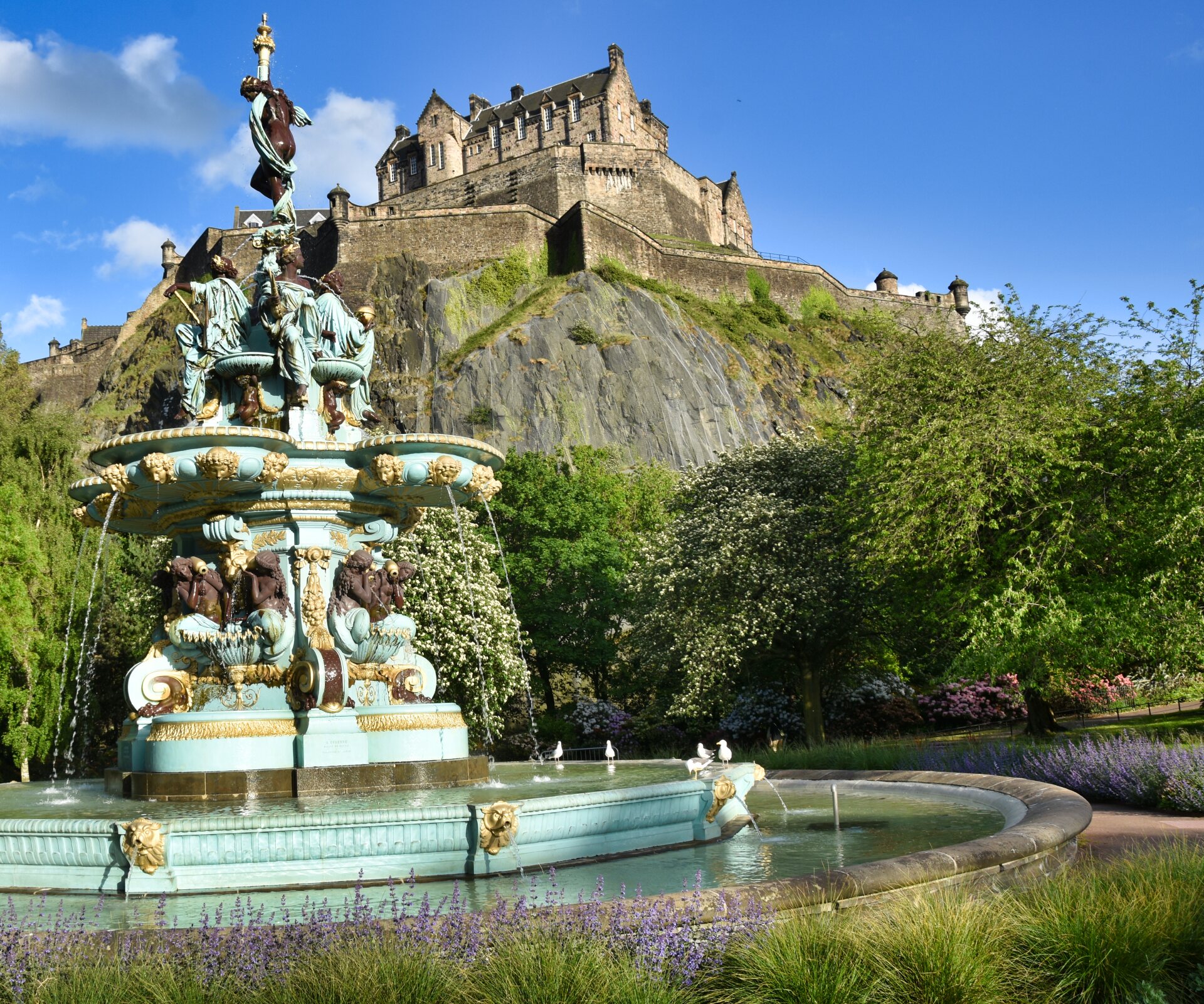 Monumentalbrunnen in hellem Türkis steht in einem Park. Im Hintergrund thront Edinburgh Castle auf einem steilen Felsen.