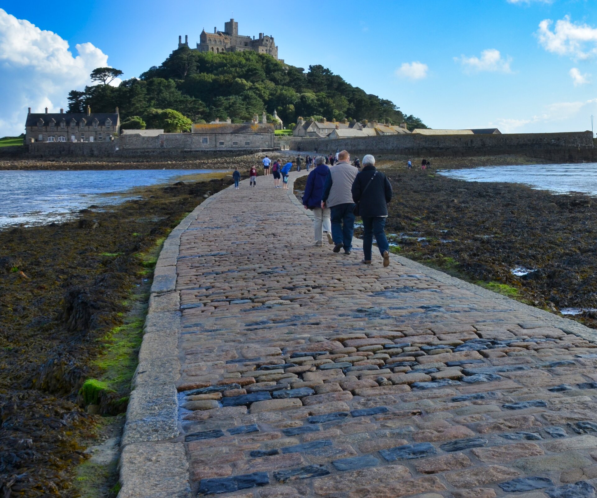 Drei Personen laufen auf einem Causeway, einer Straße aus Kopfsteinpflaster durch das Meer auf einen Berg zu.