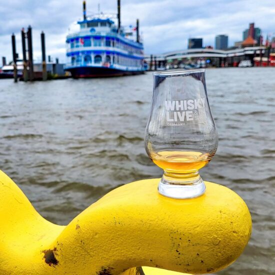 Glas der Whisky Live Germany auf einem gelben Poller der Landungsbrücken Hamburg mit Schaufelraddampfer im Hintergrund.